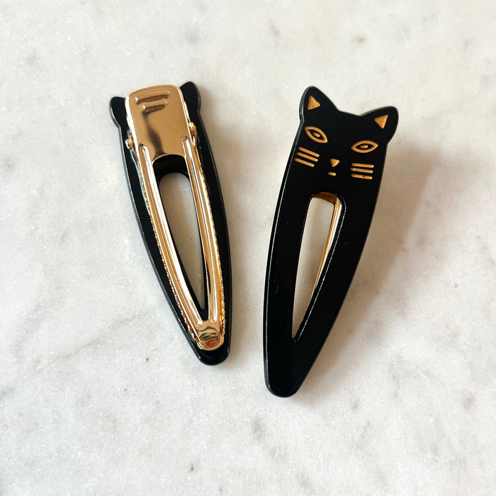 Lotta Cat Duo Clips in Black showing front and back — black cellulose acetate cat-face snap clip with gold-etched features alongside its gold metal clip mechanism, on a marble surface
