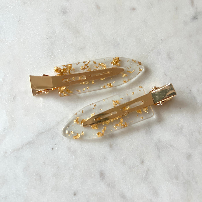 Two gold hair clips with gold flakes on a marble surface