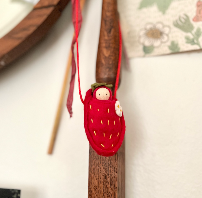 Fairy Shadow Strawberry Baby Necklace in light skin tone, hanging from a wooden dowel against a white background, showing a light-skin velour baby doll peeking out of a felted red strawberry pouch with embroidered gold seed details, a white blossom accent, green felt leaf top, and red ribbon cord