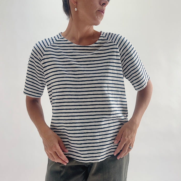 Front view of model wearing a navy and white striped boxy raglan tee with short sleeves and a relaxed fit, hands in pockets, styled with olive green pants against a white background.