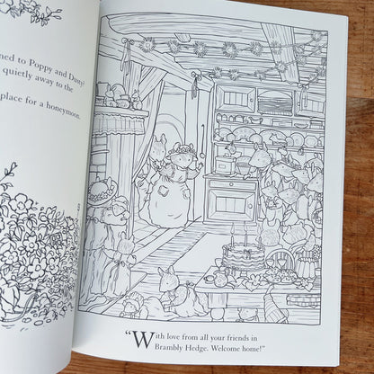 Open interior spread of the Brambly Hedge Nature Coloring Book showing a full-page uncolored line-art illustration of a mouse wedding celebration in a cozy cottage kitchen: a bride mouse in a floral headpiece greets guests at the door while other mice crowd around a table set with a tiered cake, teacups, and food, with shelves of crockery, hanging flower garlands, and a quote reading "With love from all your friends in Brambly Hedge. Welcome home!" below, displayed on a warm wood surface.