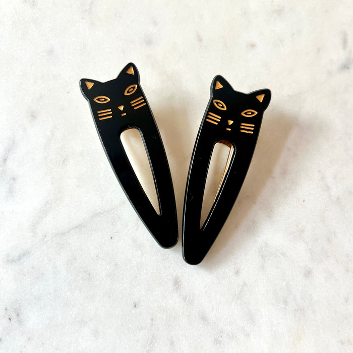 Lotta Cat Duo Clips in Black — a pair of black cellulose acetate snap clips with gold-etched cat-face tops including eyes, ears, whiskers, and nose, on a marble surface