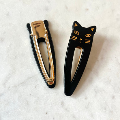 Lotta Cat Duo Clips in Black showing front and back — black cellulose acetate cat-face snap clip with gold-etched features alongside its gold metal clip mechanism, on a marble surface