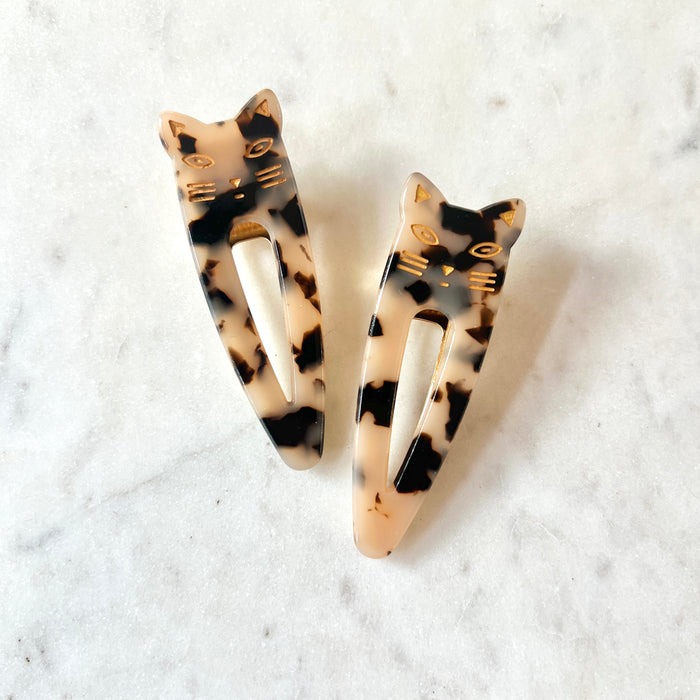 Lotta Cat Duo Clips in Light Tortoise — a pair of cream and black speckled cellulose acetate snap clips with gold-etched cat-face tops, on a marble surface