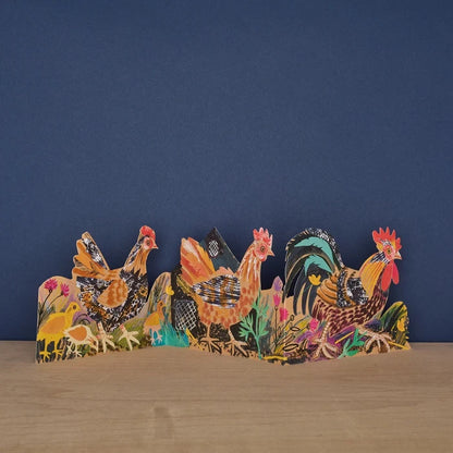 Decorative paper cutout card of chickens on a wooden surface with a blue background