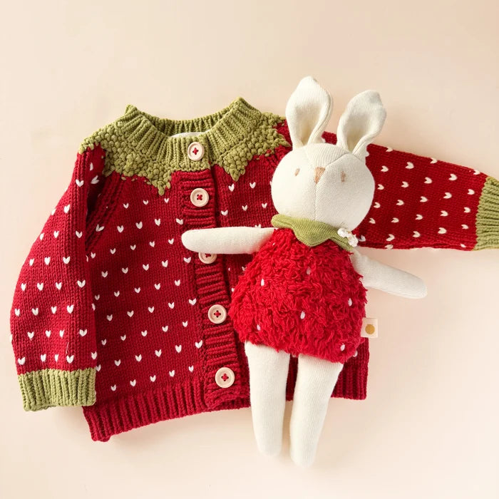 Red cotton knit baby cardigan with white heart pattern, olive green yoke and cuffs, and pink buttons, styled flat with a coordinating bunny plush toy.