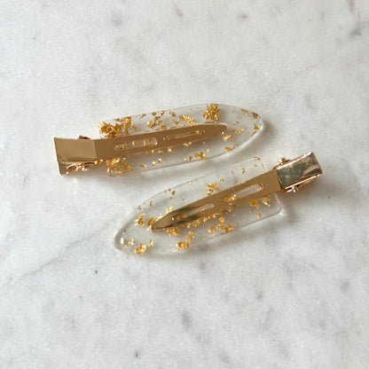 Two gold hair clips with gold flakes on a marble surface