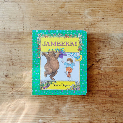 Front cover of Jamberry board book by Bruce Degen, featuring a bright green background scattered with iridescent blue and purple berry dots and a floral vine border of strawberries and white blossoms. A central framed panel on a light blue background shows a large brown bear rearing up on its hind legs and tossing a purple hat full of blueberries into the air, while a small red-haired boy in a blue shirt and orange trousers reaches up with a yellow hat to catch them. The title is printed in bold purple seri
