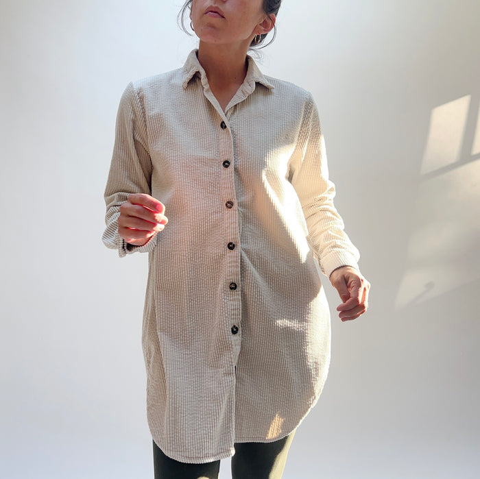 Model wearing Cut Loose Long Lux Corduroy Shirt in Moonbeam, full front view in warm dappled sunlight against white wall showing pale ecru wide-wale 100% cotton corduroy, classic point collar, dark tortoiseshell button-front placket, long sleeves, and relaxed longline silhouette hitting mid-thigh with curved hem, styled with dark slim trousers and gold hoop earrings