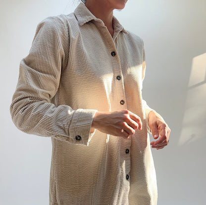 Model wearing Cut Loose Long Lux Corduroy Shirt in Moonbeam, close-up front detail in warm dappled sunlight showing pale ecru wide-wale 100% cotton corduroy texture, classic point collar open at neckline, dark tortoiseshell button-front placket, and long sleeves with button cuffs, styled with gold rings and bracelet