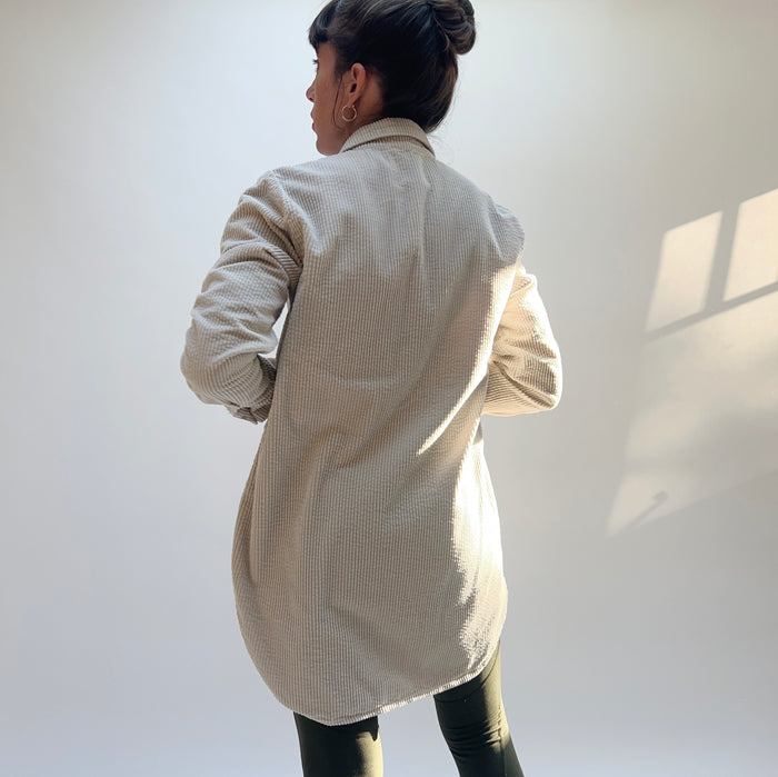 Model wearing Cut Loose Long Lux Corduroy Shirt in Moonbeam, full back view in warm dappled sunlight against white wall showing pale ecru wide-wale 100% cotton corduroy, relaxed longline silhouette hitting mid-thigh, long sleeves with button cuffs, and curved hem, styled with dark slim trousers and gold hoop earrings