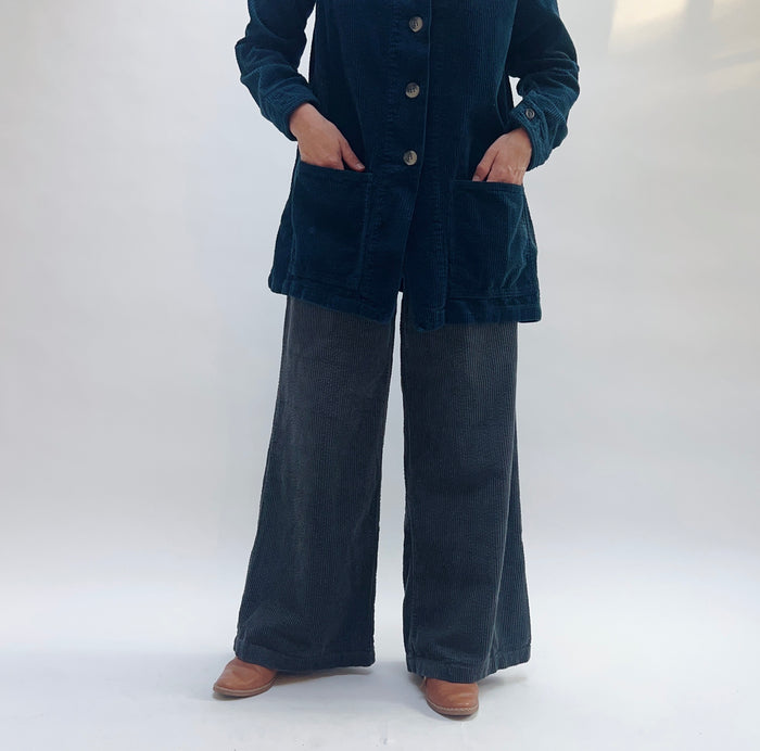 Model wearing Cut Loose Lux Corduroy Wide Leg Zip Pants in Gull, full front view against white studio backdrop showing deep slate blue-grey wide-wale corduroy, high-rise waist, very wide sweeping leg falling to ankle, styled with matching teal Cut Loose lux corduroy chore jacket with patch pockets and tan leather cowboy boots, hands in jacket pockets