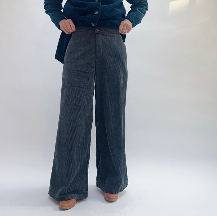 Model wearing Cut Loose Lux Corduroy Wide Leg Zip Pants in Gull, front close-up view against white studio backdrop showing deep slate blue-grey wide-wale corduroy, high-rise zip and button fly waistband with hands pulling open jacket, dramatically wide sweeping leg to ankle, styled with teal Cut Loose corduroy chore jacket and tan leather cowboy boots