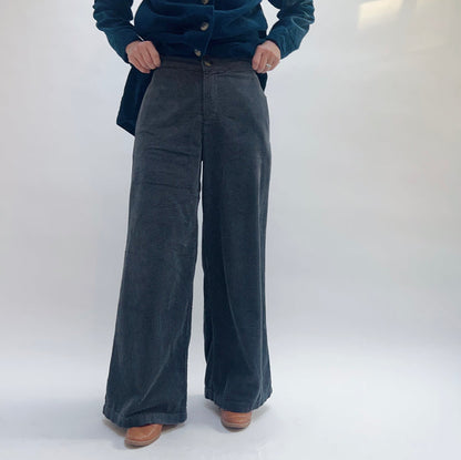 Model wearing Cut Loose Lux Corduroy Wide Leg Zip Pants in Gull, front close-up view against white studio backdrop showing deep slate blue-grey wide-wale corduroy, high-rise zip and button fly waistband with hands pulling open jacket, dramatically wide sweeping leg to ankle, styled with teal Cut Loose corduroy chore jacket and tan leather cowboy boots