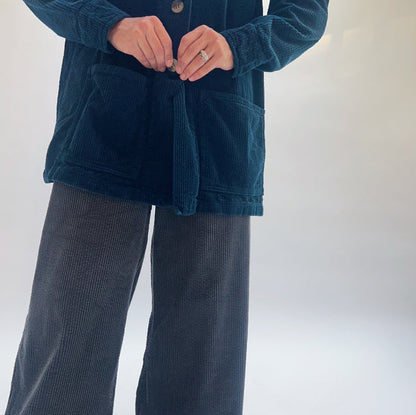 Close-up detail of Cut Loose Lux Corduroy Wide Leg Zip Pants in Gull at hip level showing deep slate blue-grey wide-wale corduroy texture, high-rise waistband, and wide leg opening, alongside the hem of a teal Cut Loose lux corduroy chore jacket with patch pockets, model wearing stacked silver rings