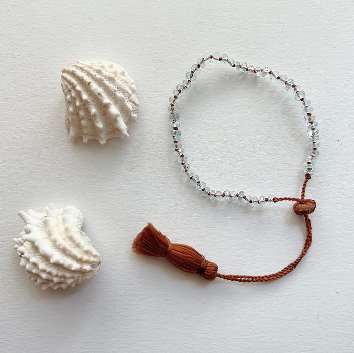 An aquamarine beaded bracelet with a waxed silk cord and an adjustable tassel closure, displayed next to two seashells.