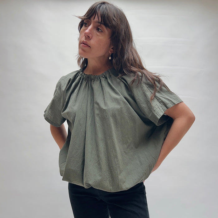 Model wearing the Uzi Nina Top in Army, three-quarter front view looking up, voluminous army green cotton blouse with gathered ruched neckline and wide cap sleeves, styled with black pants