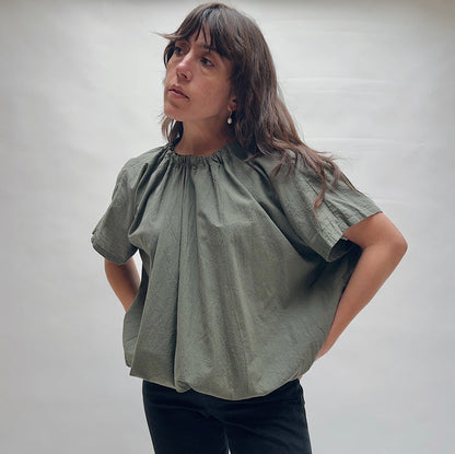 Model wearing the Uzi Nina Top in Army, three-quarter front view looking up, voluminous army green cotton blouse with gathered ruched neckline and wide cap sleeves, styled with black pants