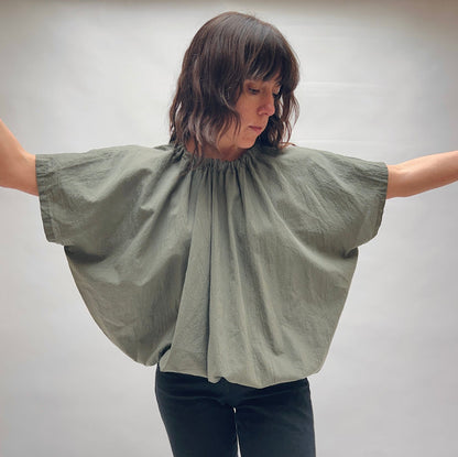 Model wearing the Uzi Nina Top in Army, full-length front view with arms extended wide, showing dramatic voluminous silhouette and gathered ruched neckline of army green finely woven cotton blouse, styled with black pants
