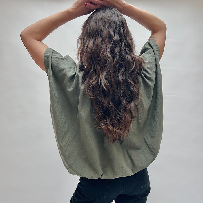 Model wearing the Uzi Nina Top in Army, back view with hands in hair, showing wide draped back and cap sleeve seam detail of voluminous army green cotton blouse with subtle wrinkle texture, styled with black pants