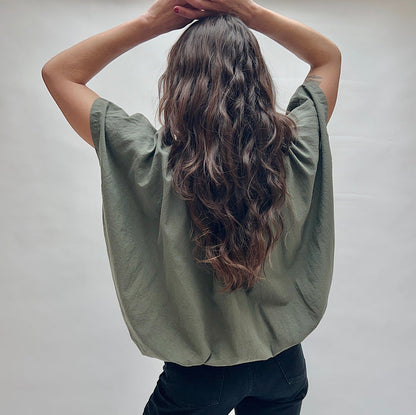 Model wearing the Uzi Nina Top in Army, back view with hands in hair, showing wide draped back and cap sleeve seam detail of voluminous army green cotton blouse with subtle wrinkle texture, styled with black pants