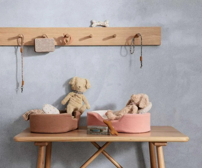 Maileg Plush Dog in Soft Sand styled in a Maileg dog-themed scene — two soft plush dogs sitting in terracotta and pink oval dog beds on a wooden bench, with miniature leashes, a toy bone, and a suitcase, against a grey wall with a wooden peg rail