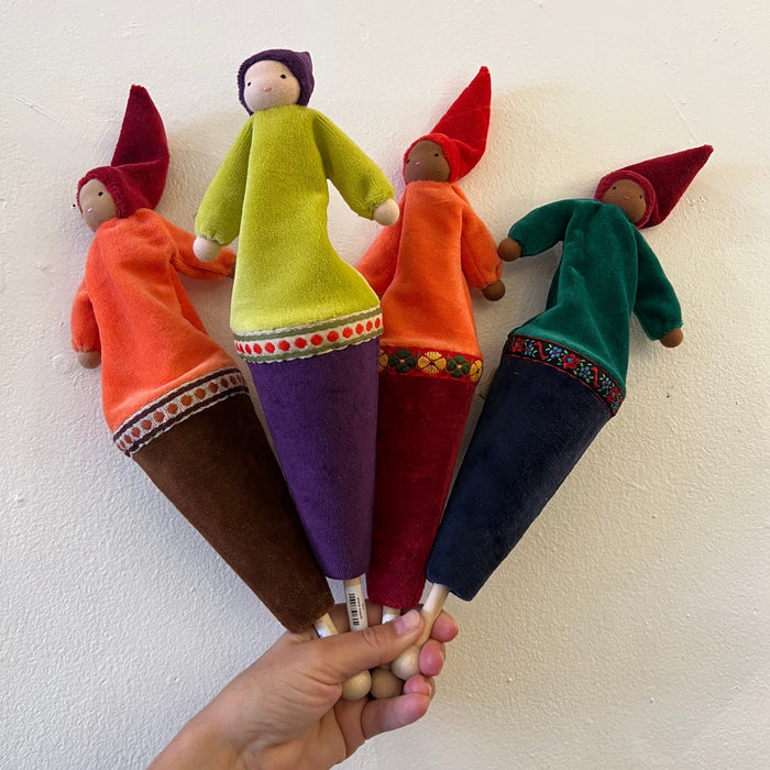 Fairy Shadow Popup Puppets, four styles fanned out and held in a hand against a white textured wall, showing cotton velour gnome-style dolls with pointed hats in various outfit colorways — orange and brown cone, yellow-green and purple cone, orange and red cone, and teal and navy cone — each with decorative ribbon trim and a wooden push stick, representing light and dark skin tone variants