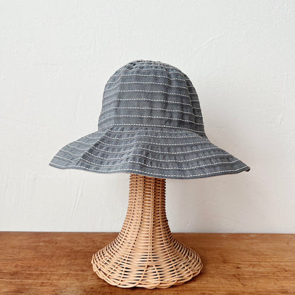 Sage ribbon bucket hat with white stitch stripe detail and wide brim, shown on a wicker hat stand — front view