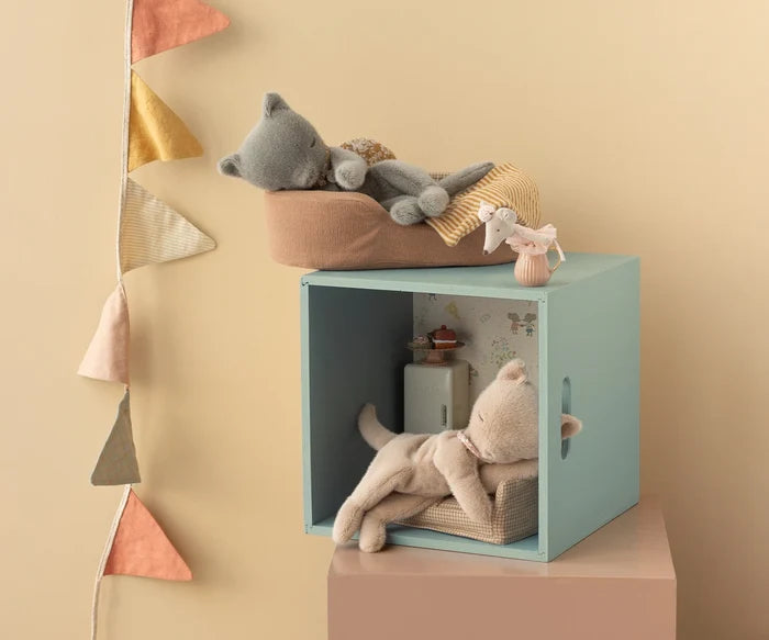Maileg Small Plush Sleeping Kitten in Cream Peach styled in a Maileg room scene — a cream-peach sleeping kitten resting inside a blue wooden cat house with a wicker basket bed, beside a grey Maileg kitten sleeping in a linen cat bed on top, with a paper bunting garland in the background