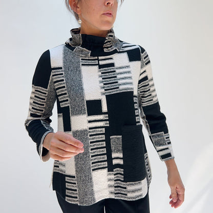 Model wearing the Bryn Walker Square One Pullover in Dove, angled front view showing mixed jacquard geometric patchwork print in black, white, and grey, ruched wired collar, large center front patch pocket, 3/4 sleeves, and relaxed A-line silhouette, styled with black pants