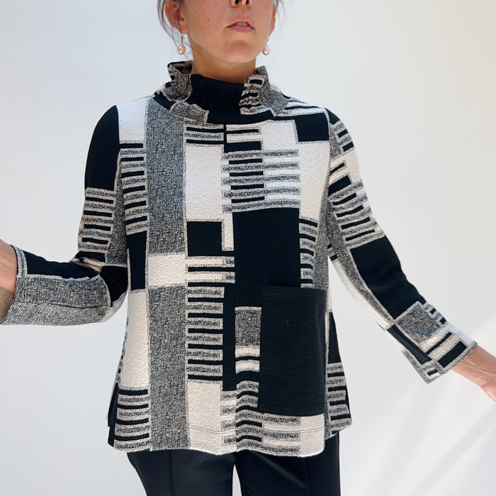 Model wearing the Bryn Walker Square One Pullover in Dove, full front view showing mixed jacquard geometric and stripe patchwork print in black, white, and grey, ruched wired collar, large center front patch pocket, 3/4 sleeves, and curved tunic hem, styled with black pants