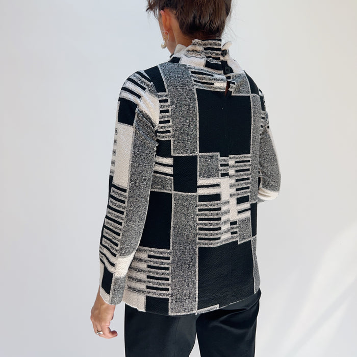Back view of model wearing the Bryn Walker Square One Pullover in Dove, showing mixed jacquard geometric patchwork print in black, white, and grey, ruched wired collar, 3/4 sleeves, side slits, and relaxed A-line silhouette, styled with black pants